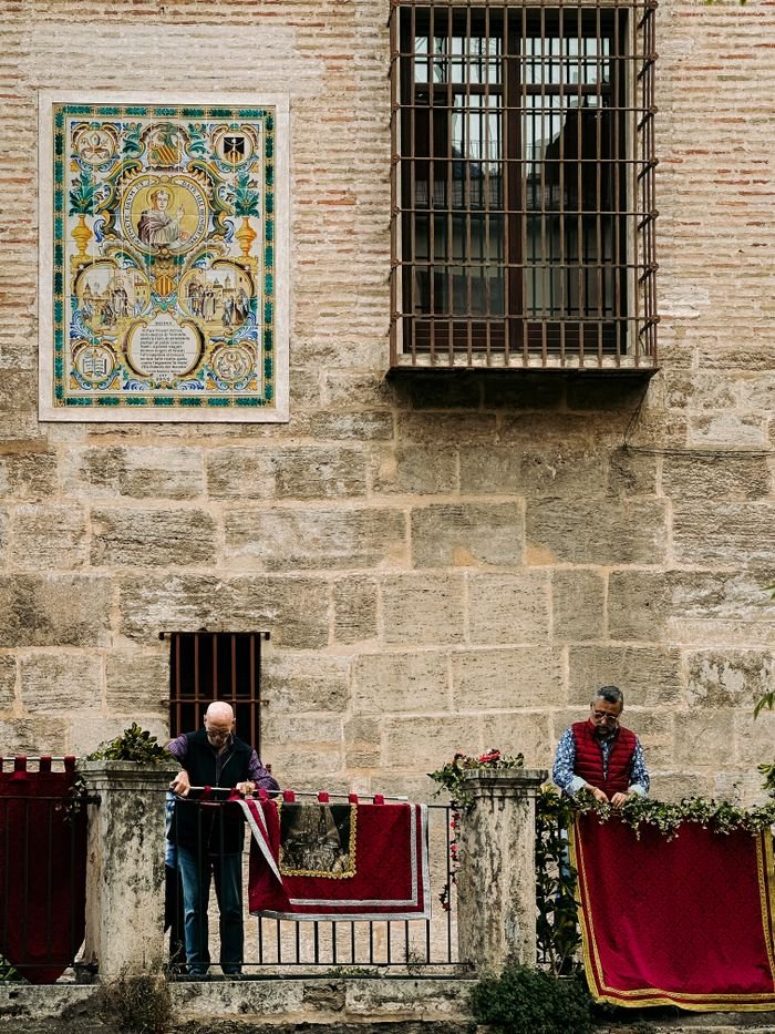 Home Two men decorate a historic building with red fabric in Valencia, Spain, showcasing traditional art and culture.